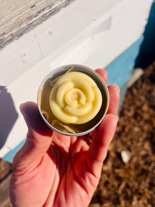 Hand holding a small metal container with a rose-shaped lotion bar against a blurred outdoor background
